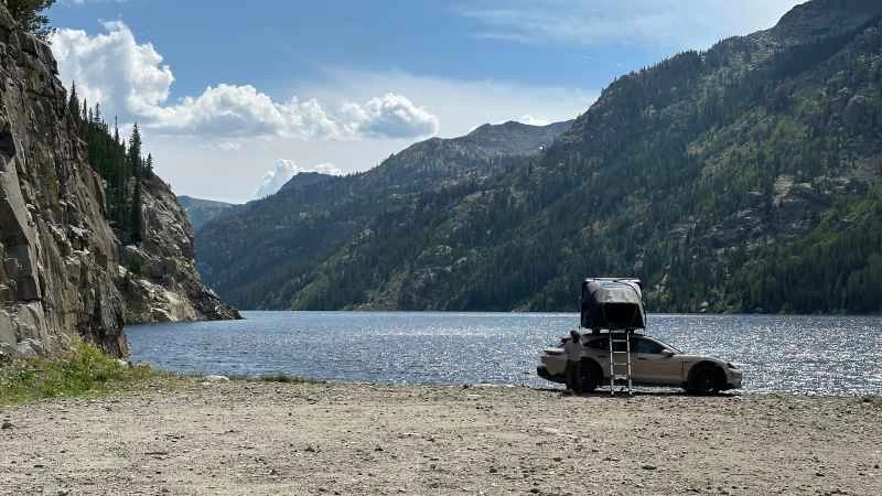 一輛SUV，車頂上架著一個簡易帳篷，旁邊放著一個梯子，停在山間湖泊前。