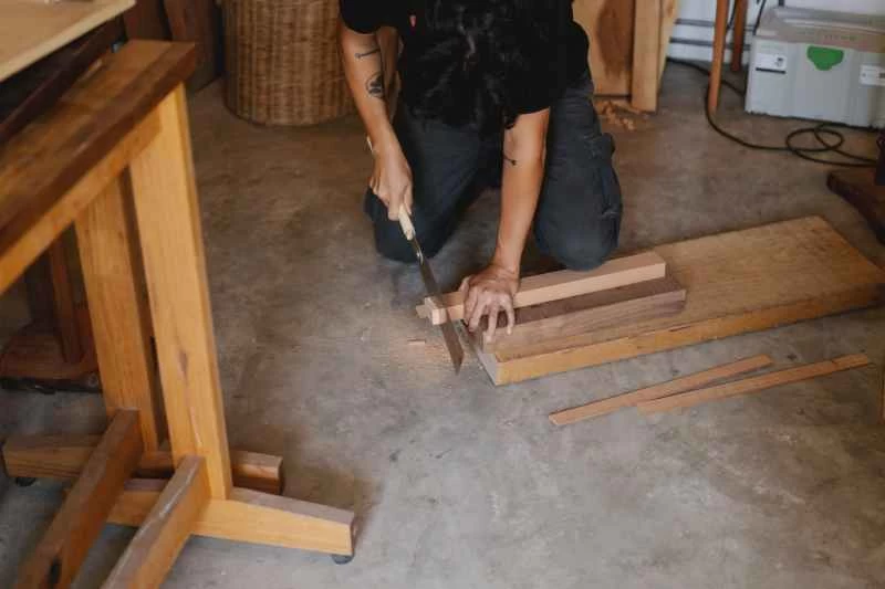 一名男子跪在木工車間的地板上，用手鋸鋸斷一小塊木頭。