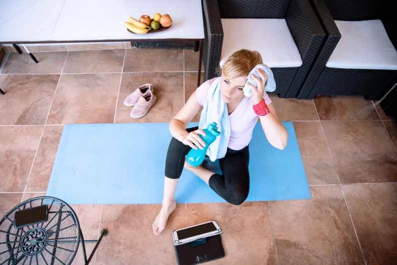 一個綁著紅色馬尾辮、穿著粉紅色襯衫和黑色運動緊身褲的人正在藍色瑜珈墊上喝水和擦汗。