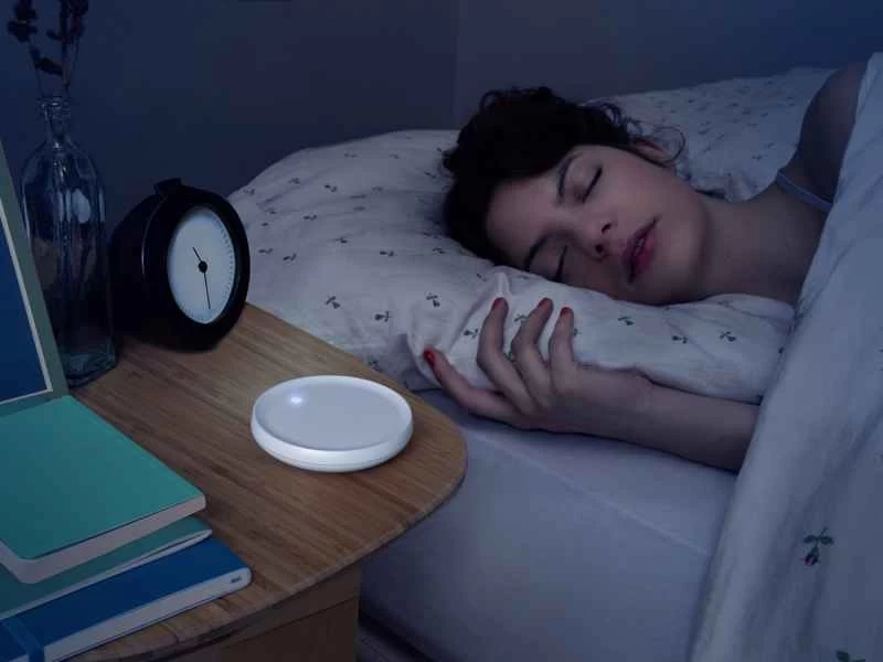 一位女士睡在床上，床頭櫃上放著一台 Dodow 助眠設備。